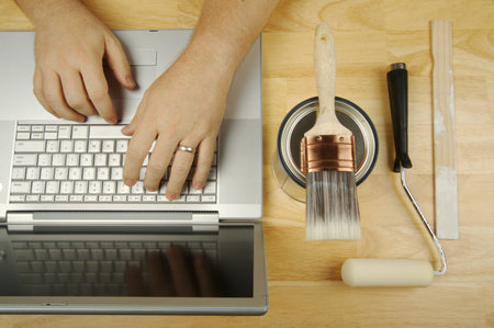 Handyman Researches on Laptop with paint brush, roller and wood stir stick by his side. Great image...