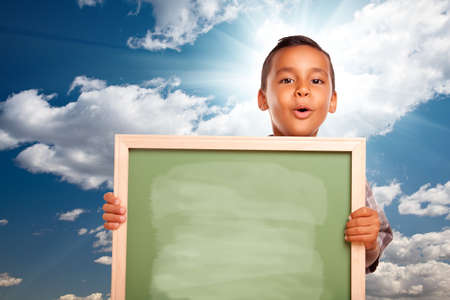 Proud Hispanic Boy Holding Blank Chalkboard Over Blue Sky and Clouds with Sun Burst.の写真素材