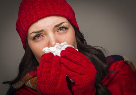 Sick Mixed Race Woman Wearing Winter Hat and Gloves Blowing Her Sore Nose with a Tissue.の写真素材