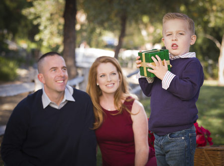 Handsome Young Boy Holding a Christmas Gift in the Park While His Mom and Dad Look On.の写真素材