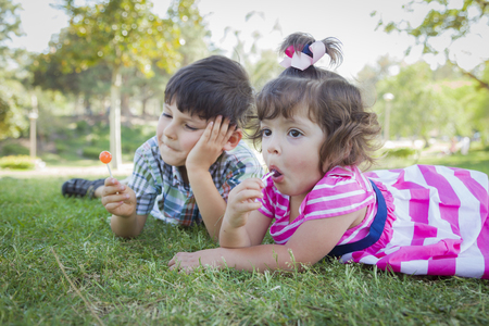 Cute Young Brother and Baby Sister Enjoying Their Lollipops Outdoors on the Grass.の写真素材