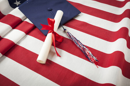 Graduation Cap with Tassel and Red Ribbon Wrapped Diploma Resting on American Flag の写真素材