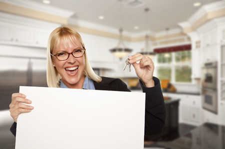 Happy Young Woman Holding Blank Sign and Keys Inside Beautiful Custom Kitchen.の写真素材