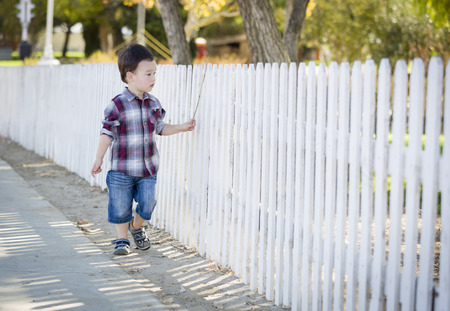 Cute Young Mixed Race Boy Walking with Stick Along White Fence.の写真素材