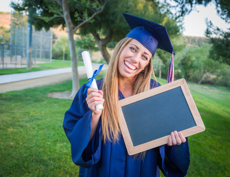 Excited Young Woman Holding Diploma and Blank Chalkboard Wearing Cap and Gown Outdoors.