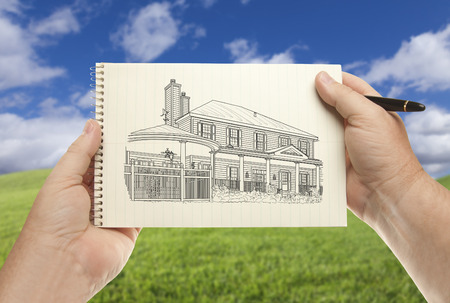 Male Hands Holding Pen and Pad of Paper With House Drawing Over Empty Grass Field and Sky.の写真素材