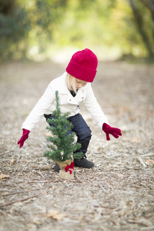 Baby Girl In Red Mittens and Cap Near Small Christmas Tree Outdoors.の写真素材