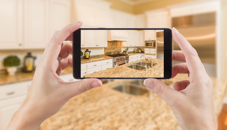 Female Hands Holding Smart Phone Displaying Photo of Kitchen Behind.の写真素材