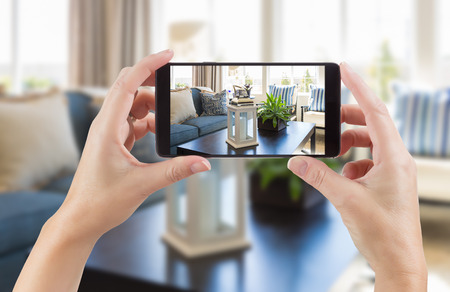 Female Hands Holding Smart Phone Displaying Photo of House Interior Living Room Behind.の写真素材