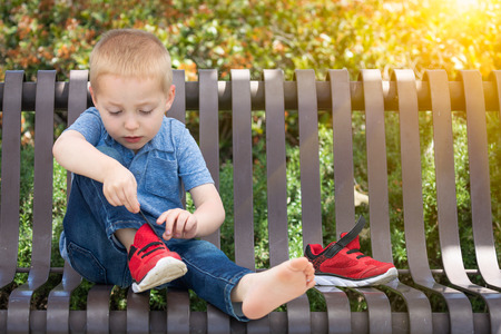 Young Boy Sitting On A Bench Putting On His Shoes At The Park.の写真素材