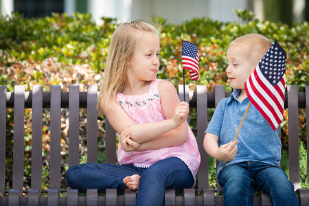Young Sister and Brother Comparing Each Others American Flag Size On The Bench At The Park.の写真素材