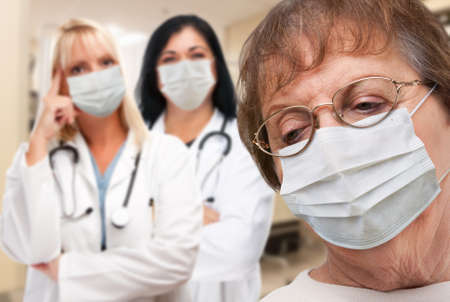 Senior Adult Female Looking Down as Doctors stand Behind All Wearing Medical Face Masks.の写真素材