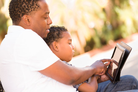 African American Father and Mixed Race Son Using Computer Tablet on Bench in Park.の写真素材