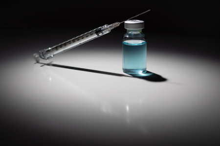 Syringe Needle and Medicine Vial With Light Blue Chemical Spot Lit on Reflective Background.