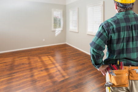 Contractor Wearing Toolbelt and Hard Hat Facing Empty Room with Hard Wood Floors.