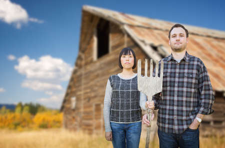 Biracial Chinese and Caucasian Young Couple Holding Wooden Pitch Fork In Front of Rustic Barn In The Country.の写真素材