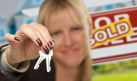 Female Real Estate Agent Presenting House Keys In Front of Sold For Sale Sign & New House. Focus is on the Keys.の写真素材