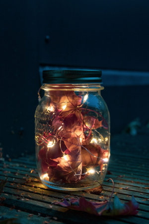Glass mason jar with LED fairy lights and dried red maple leaves on wooden patio in the dark in fall...