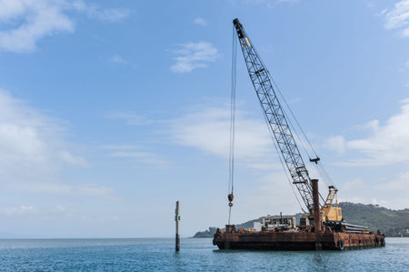 A large crane sits on a barge in a bay with hills behind it