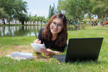 young woman working on laptop outdoors. image of female student working on laptop while lying on a grass in a park with booksの写真素材