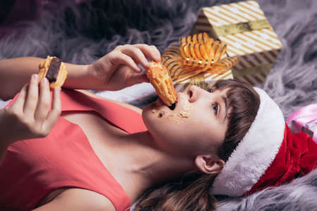 A teenage girl in a Santa hat eats Christmas cakes lying on the carpet, in the background golden boxes of gifts. High quality photoの写真素材