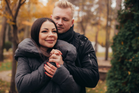 A man hugs and holds the hand of his girlfriend as she smiles in the cold weather in the autumn park. High quality photoの写真素材