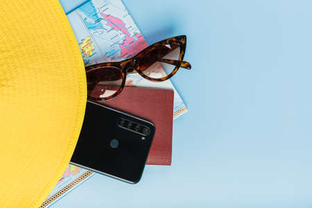 Travel flatlay, world map, passport, mobile phone and sunglasses lie under a yellow hat on a blue backgroundの写真素材