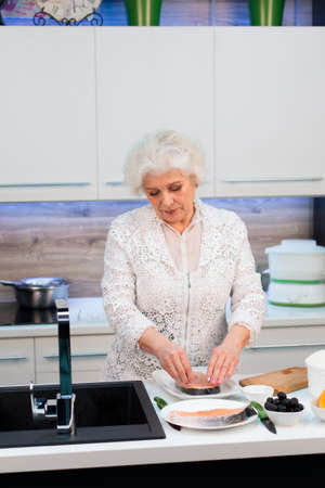 An old well-groomed blonde woman cooks a trout steak in the kitchenの写真素材