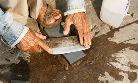 Old Laotian man sharpening a knife with his hands on a wet stone on the side of a road in Suvanakhet, Laos, Southeast Asiaの写真素材