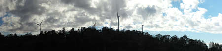 panoramic views of multiple windmills at a modern wind farm built on a green hill on a sunny blue...