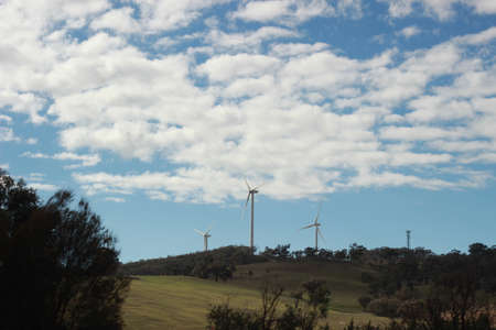 panoramic views of multiple windmills at a modern wind farm built on a green hill on a sunny blue...