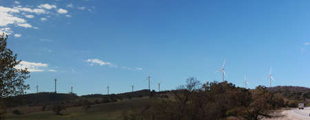 panoramic views of multiple windmills at a modern wind farm built on a green hill on a sunny blue...