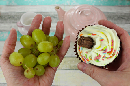 Healthy grapes or cupcake? Hands holding a healthy snack of grapes in one hand and an unhealthy cupcake in the other. Tea items in background.の写真素材