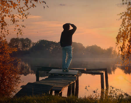 a man watches a picturesque sunset on the shore of a golden warm autumn lakeの写真素材