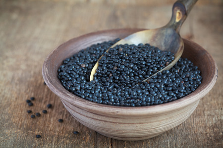 Dry black lentil beluga in a clay bowl and in a scoop on wooden table, selective focus - some beans in focus, some are notの写真素材