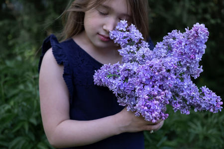 Cute little girl smelling lilac. Person holding flowers on dark natural background. Child enjoying bouquet outside. Going to park, forest in summer and spring. Connection with nature ideaの写真素材