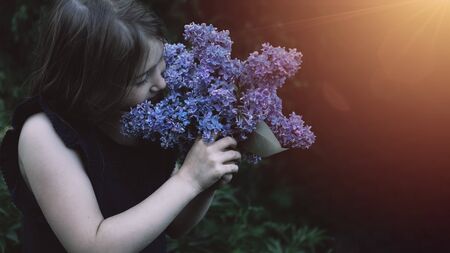 Cute little girl smelling lilac. Person holding flowers on dark natural background. Child enjoying bouquet outside. Going to park, forest in summer and spring. Connection with nature ideaの写真素材