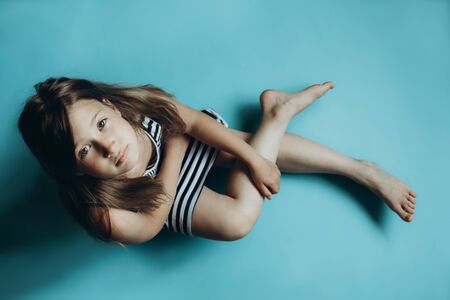 Girl sitting on plain blue background looking at camera. Child glancing up top view. Simple wallpaper, background. Serious and sad facial expression. Kid in striped dress with legs crossedの写真素材