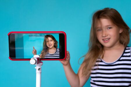 Girl posing in front of phone camera for selfie. Focus o smartphone screen fixed on tripod. Recording homemade video. Happy smiling child. Cute kid pointing up. Simple wallpaper, backgroundの写真素材