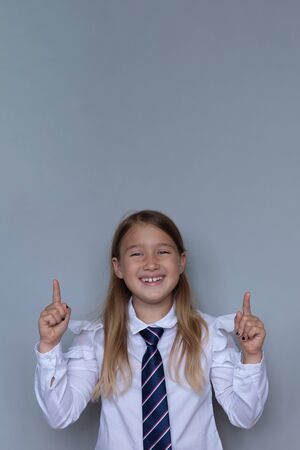 Young schoolchild pointing up portrait. Preteen schoolgirl looking at camera, gesturing at ceiling. Happy girl, kid in school uniform. Back to school, joyful, cheerful pupil, student ready for classの写真素材