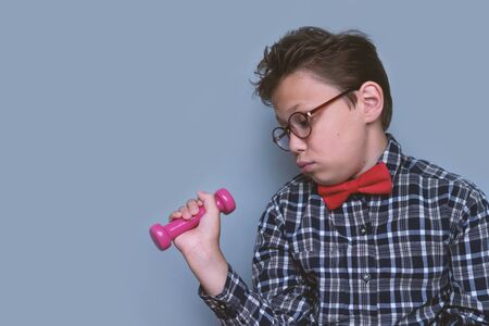 Smart boy with glasses in checkered shirt and red bow tie looks at the dumbbell in bewilderment. Dislike sport and healthy life concept. Nerd and sportの写真素材