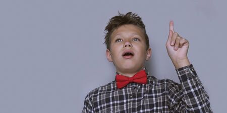 Young schoolchild pointing up portrait. Preteen schoolboy gesturing at ceiling. Happy boy, kid incheckered shirt with red bow tie. Back to school, cheerful pupil. I have an idea and insight conceptの写真素材
