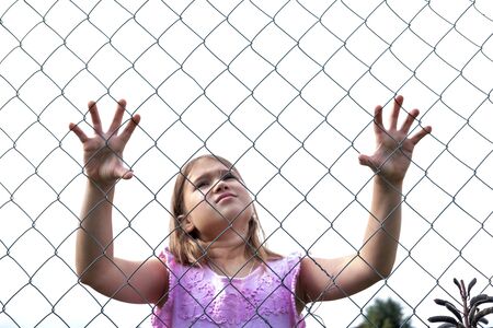 Sad girl behind metal wire mesh fence. Abandoned upset and lonely child outdoor. Depressed preteen...