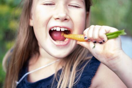 Girl eating carrot. Child face closeup. Healthy, organic food. Fresh ripe vegetable. Summertime, spring sweet treat. Eco quick snack. Natural handpicked tasty food. Vegetarian and vegan dietの写真素材