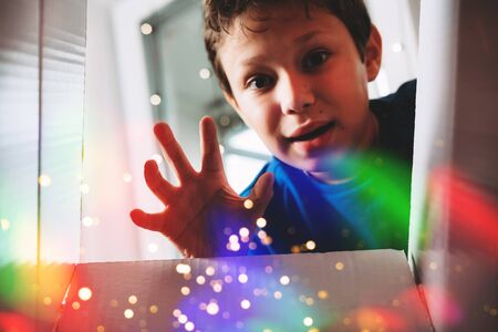 Happy boy unwrapping present. Child excited with Christmas holiday gift concept. Kid looking inside magic box with sparkles and rainbow bokeh lights effect. Teenager with getting birthday surpriseの写真素材