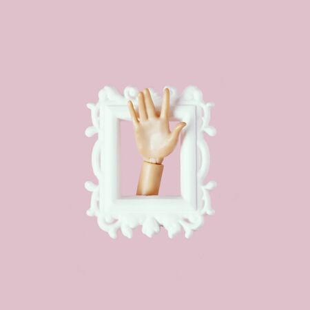 Doll hand in picture frame illustration. Plastic toy arm in ornate white border isolated on soft pink color background. Creative childish composition, minimalistic style design elementの写真素材