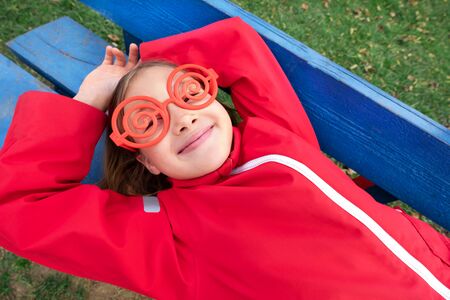 Girl with sunglasses and red jacket lies on park bench and smile. Spending time outdoors. Child playing outside in autumn. Childhood memories. Going on journey, adventure in wilderness.の写真素材