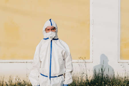 Man wearing protective COVID costume, mask, respirator and gloves. Doctor stay and looking forward on yellow wall background. Pandemic, self-isolation and quarantine finish concept.の写真素材