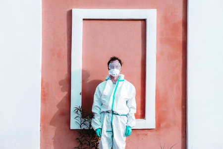 Man wearing protective COVID costume, mask, respirator and gloves. Doctor stay and looking forward on pink and white wall background. Pandemic, self-isolation and quarantine finish concept.の写真素材