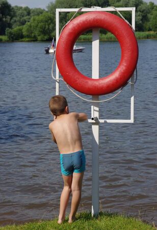 The boy is standing on the shore of the pond next to the big red life ringの写真素材
